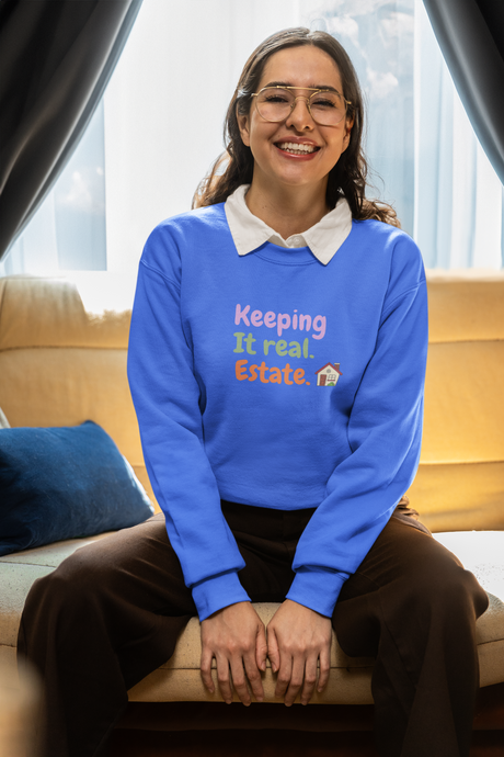 Blue unisex crewneck sweatshirt with "Keeping It Real. Estate." slogan, worn by a person sitting indoors.