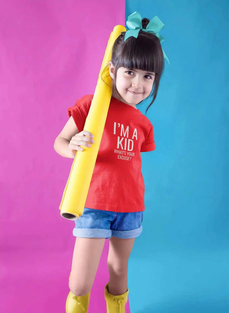 Kid wearing a red "I'm A Kid" softstyle t-shirt, holding a yellow roll, standing against a pink and blue background.