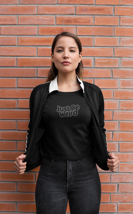 Woman wearing "Just Be Weird" dark women's cropped t-shirt against brick wall.