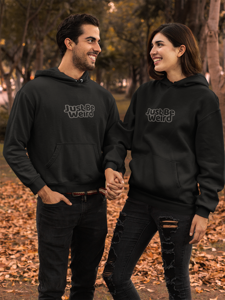 Unisex black hoodie with "Just Be Weird" text, worn by a man and woman outdoors.