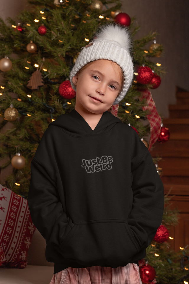 Kids wearing a "Just Be Weird" dark unisex hoodie in front of a decorated Christmas tree.