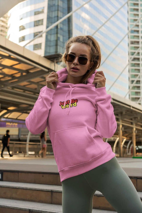 Pink hoodie with "Just Be Weird" design, worn by person outdoors against urban backdrop; Weird Unisex Heavy Blend Hoodie.